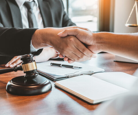 A male lawyer shaking hands with a woman at the lawyers desk. There is a gavel and notebooks with a pen.